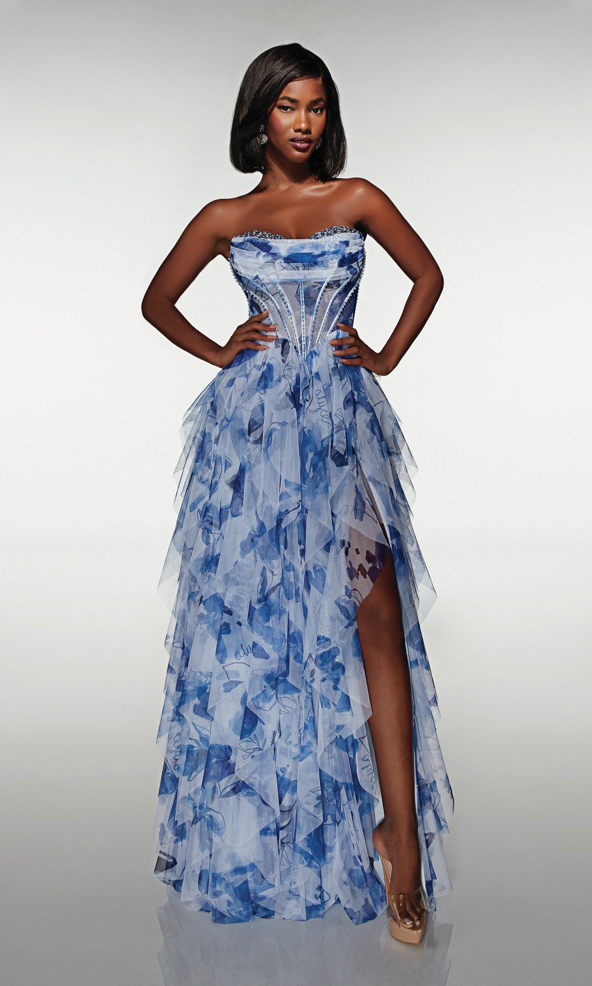A woman stands with hands on hips, modeling the Alyce 62041 long prom dress in blue/print, featuring a strapless corset bodice and layered skirt against a plain background.
